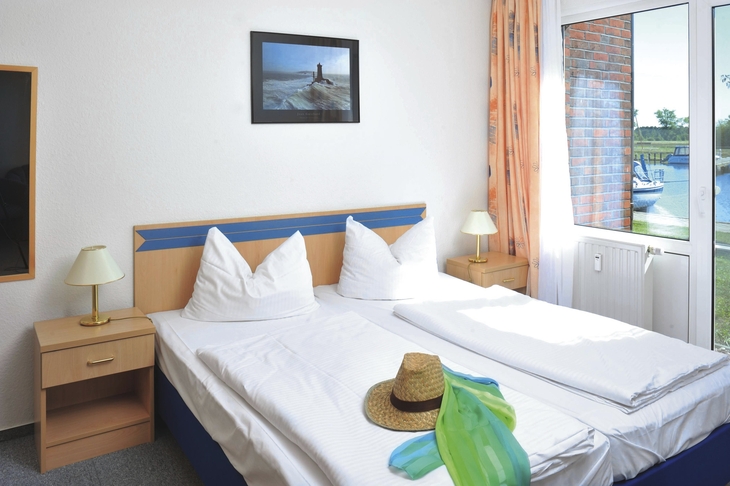 Hotelzimmer mit Doppelbett, Strohhut und Blick auf Wasser durch Fenster.