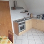 Kleine Küche mit Holzschränken, Herd, Kühlschrank und Tisch mit kariertem Tuch.