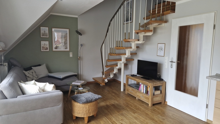 Wohnzimmer mit Sofa, Sessel, Fernseher und offener Treppe auf Holzboden.