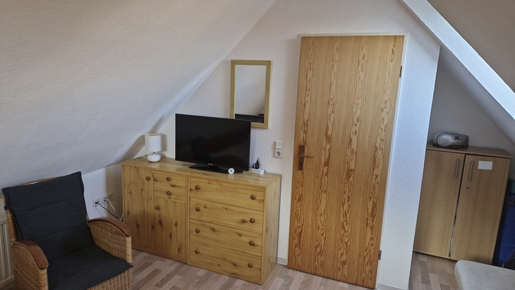 Dachzimmer mit Holzkommode, Fernseher und Tür, neben Rattanstuhl unter Dachschräge.