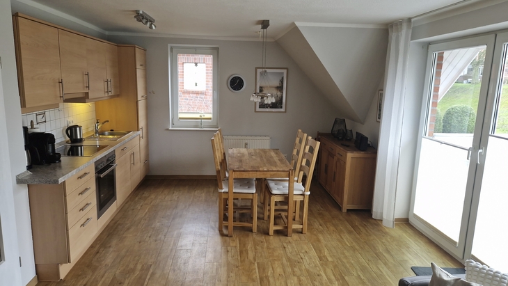 Küche und Essbereich mit Holztisch, Stühlen, Küchenzeile und großen Fenstern.