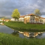Gebäude am Fluss, von Bäumen umgeben, mit Wolken und Regenbogen im Hintergrund.