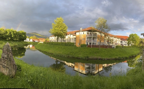 Gebäude am Fluss, von Bäumen umgeben, mit Wolken und Regenbogen im Hintergrund.