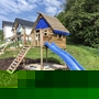 Holzspielplatz mit Kletterhaus und blauer Rutsche, umgeben von Grünflächen.