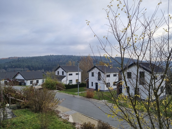 Wohnsiedlung mit weißen Häusern und bewaldeten Hügeln im Hintergrund bei bewölktem Himmel.