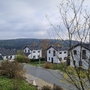 Wohnsiedlung mit weißen Häusern und bewaldeten Hügeln im Hintergrund bei bewölktem Himmel.