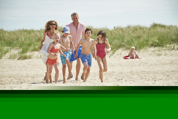 Familie spielt am sonnigen Sandstrand mit Dünen im Hintergrund.