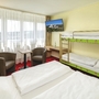 Zimmer mit Doppelbett, Etagenbett, Fernseher, zwei Sesseln und Fenster mit Vorhängen.