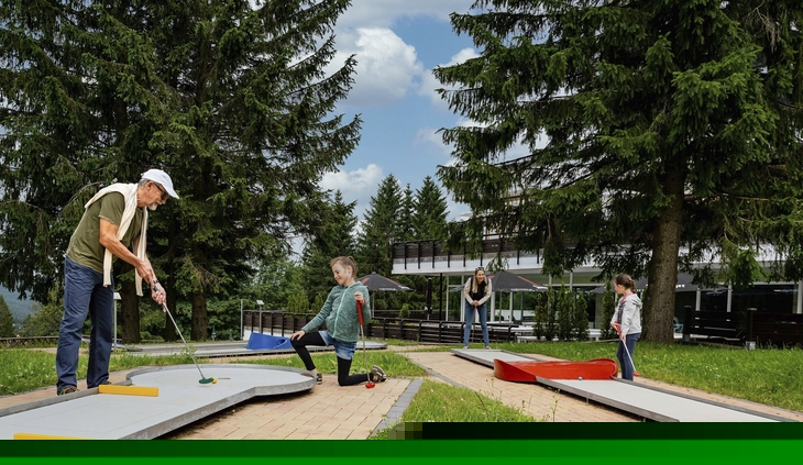Familie spielt Minigolf im Freien, umgeben von Bäumen.