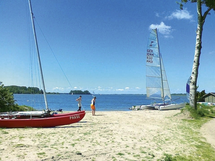 Strand mit Katamaranen und Personen am Wasser bei sonnigem Wetter.