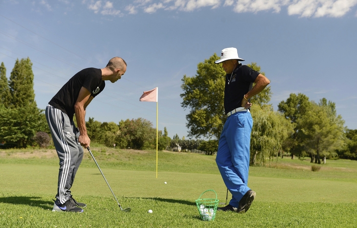 Zwei Männer beim Golfspiel auf einem Rasenplatz mit Bäumen und einem roten Flaggenstock.