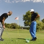 Zwei Männer beim Golfspiel auf einem Rasenplatz mit Bäumen und einem roten Flaggenstock.