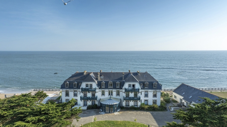 Weiße Residenz am Meer mit blauen Balkonen, umgeben von Bäumen, im Hintergrund der Ozean.