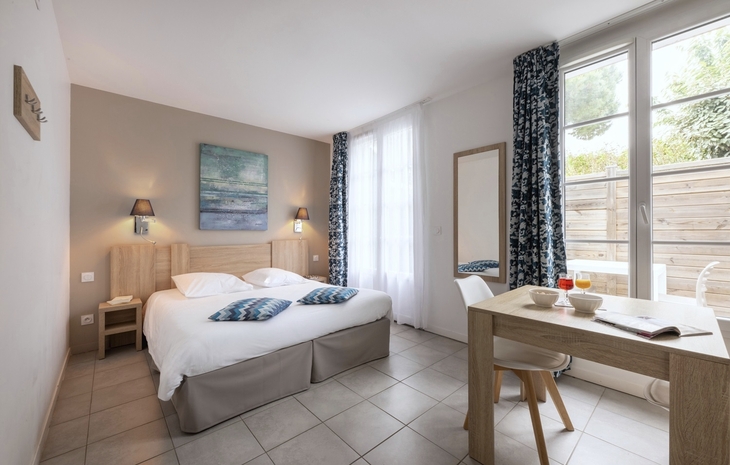 Helles Hotelzimmer mit Doppelbett, Schreibtisch und Fenster mit Vorhängen.