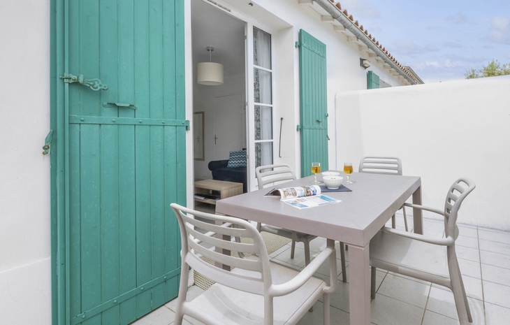 Terrasse mit Tisch, Stühlen und Blick auf Wohnzimmer durch offene Tür mit grünen Fensterläden.