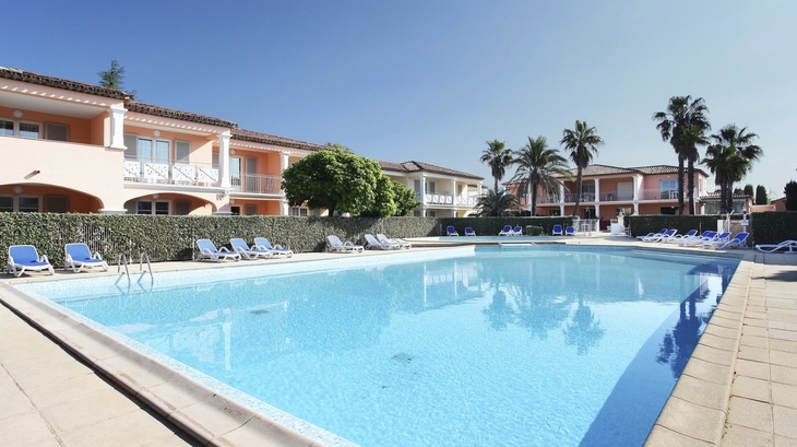 Pool vor einer Ferienanlage mit Palmen und Liegestühlen bei sonnigem Wetter.