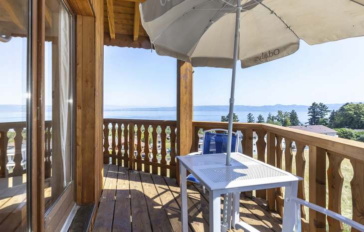 Holzbalkon mit Tisch, Stuhl und Sonnenschirm, Blick auf See und bewaldete Hügel.
