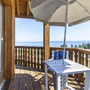 Holzbalkon mit Tisch, Stuhl und Sonnenschirm, Blick auf See und bewaldete Hügel.