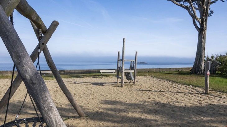 Kinderspielplatz mit Holzgeräten am Strand, Meer im Hintergrund.