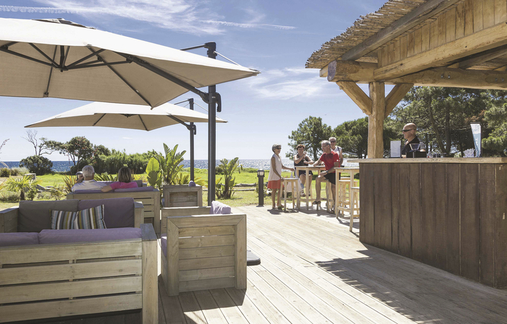Terrasse mit Sitzgelegenheiten und Sonnenschirmen neben einer Holzbar bei sonnigem Wetter.