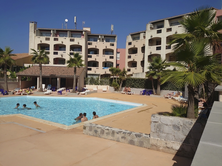 Außenansicht mit Pool, Palmen und mehrstöckigem Wohngebäude im Hintergrund.