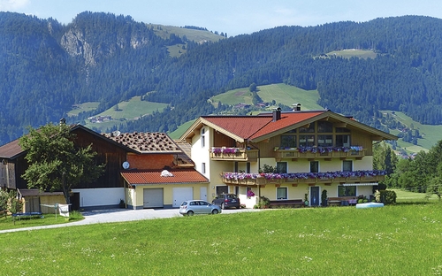 Haus in bergiger Landschaft mit blühenden Balkonen und Wiese im Vordergrund.