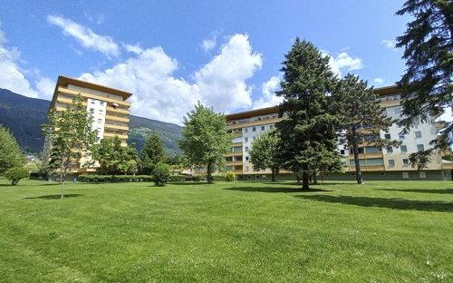 Appartementanlage mit grüner Wiese und Bäumen vor Bergen unter blauem Himmel.