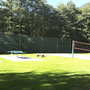 Tischtennisplatten und Volleyballnetz auf einer grünen Wiese vor Bäumen.