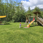 Spielplatz mit Rutsche und Kindern auf Rasenfläche vor bewaldeter Bergkulisse.