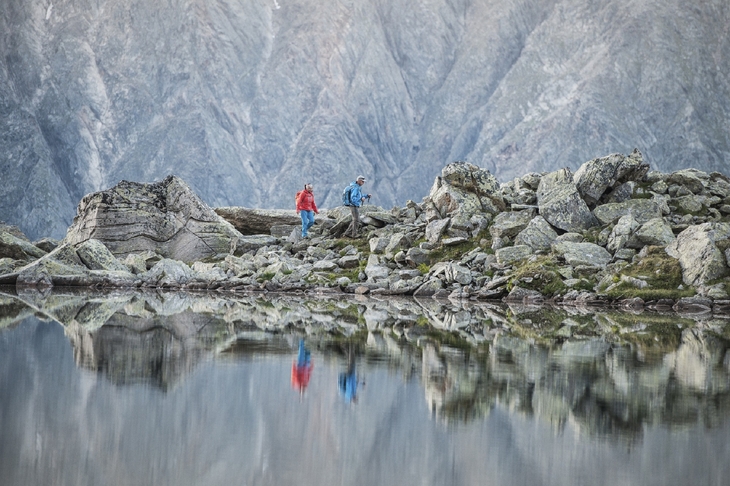Zwei Wanderer auf einem Felsweg bei einem Bergsee, Berge im Hintergrund.