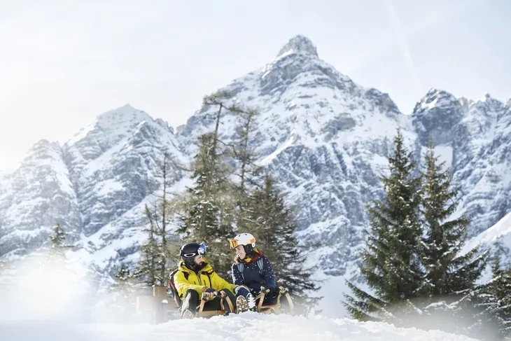 Zwei Personen rodeln im Schnee vor verschneitem Bergpanorama in Tirol.