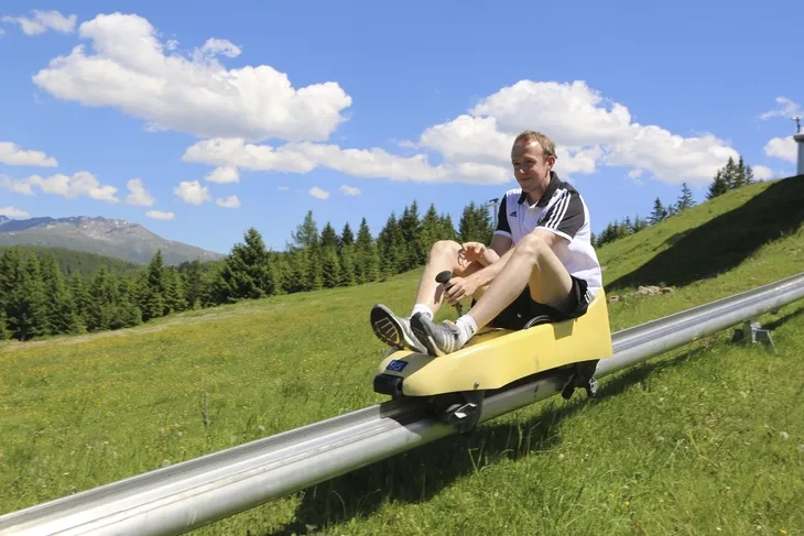 Person auf Sommerrodelbahn in grüner Alpenlandschaft mit blauem Himmel.
