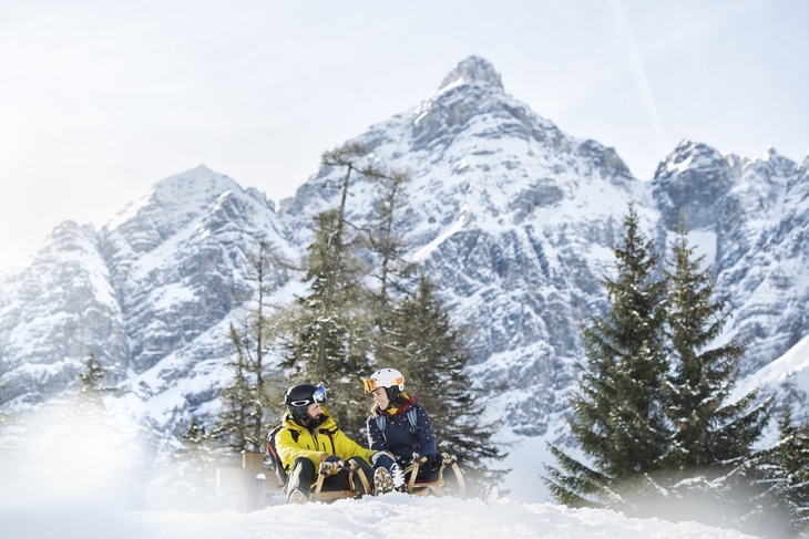 Zwei Personen rodeln im Schnee vor verschneitem Bergpanorama in Tirol.