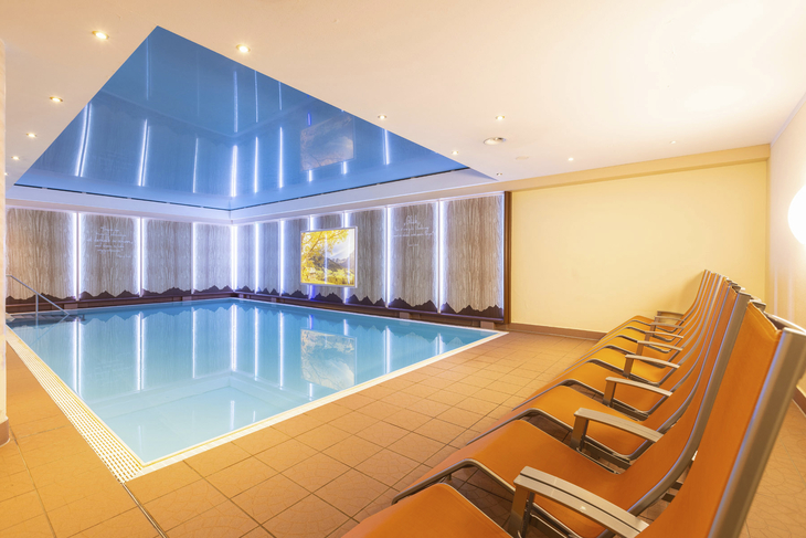 Hallenbad mit blauen Fliesen und orangefarbenen Liegestühlen im Hotel Alpenrose.