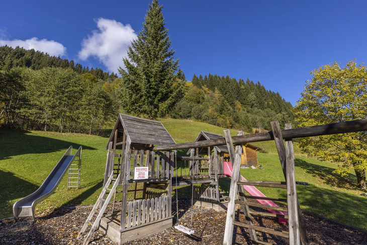 Spielplatz mit Rutsche und Klettergerüst, umgeben von Bäumen und Wiesen.