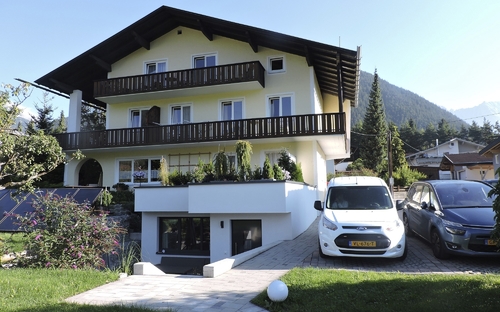Gelbes Haus mit Holzdetails, zwei Autos davor, in bergiger Umgebung.
