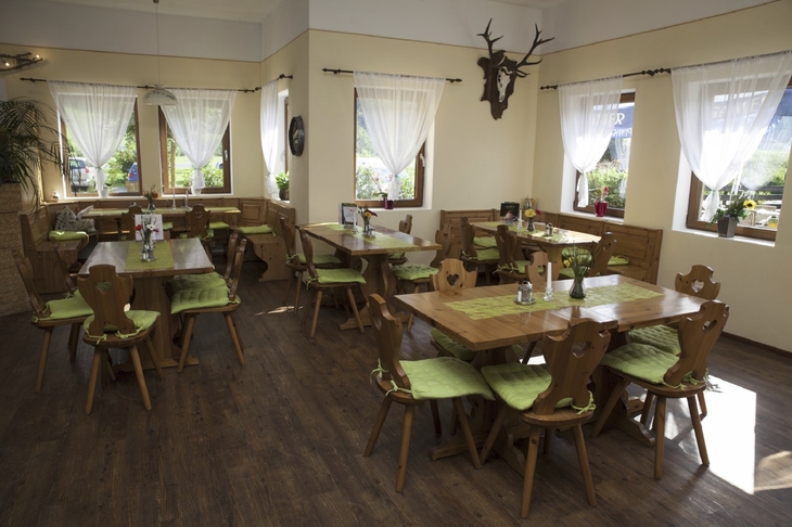 Restaurantbereich mit Holzstühlen, grünen Kissen und Vorhängen, Tageslicht durch Fenster.
