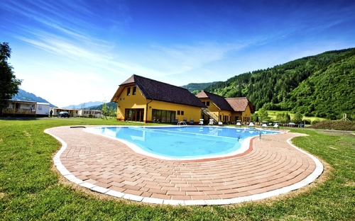 Pool vor gelben Gebäuden auf einem Campingplatz in Hügel- und Waldlandschaft.