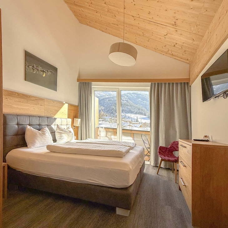 Helles Hotelzimmer mit Doppelbett, Holzmöbeln und großer Fensterfront mit Bergblick.