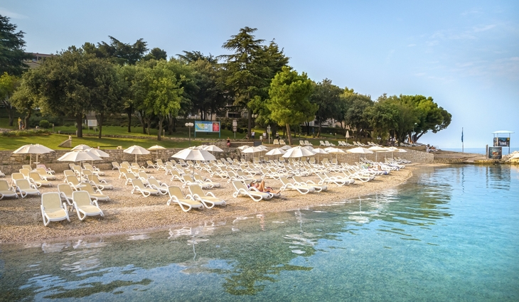 Strand mit Sonnenliegen und Sonnenschirmen am klaren Wasser, umgeben von Bäumen.