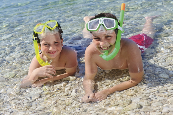 Zwei Kinder mit Schnorchelausrüstung liegen lächelnd im flachen Wasser.