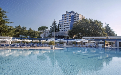 Hotelpool mit Liegestühlen, Bäumen im Hintergrund und einem Gebäudekomplex.
