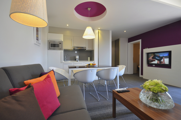 Modernes Wohnzimmer mit Küchenzeile, bunten Kissen und Blumenarrangement.