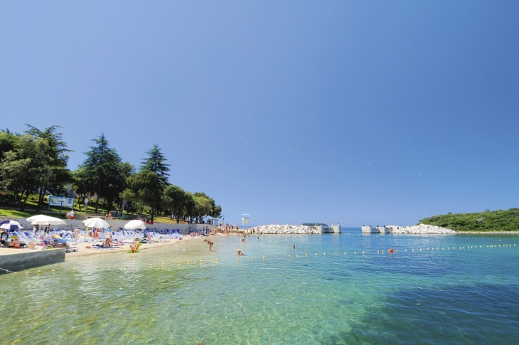 Strand mit Badegästen, Bäumen und klarem blauem Wasser unter klarem Himmel.