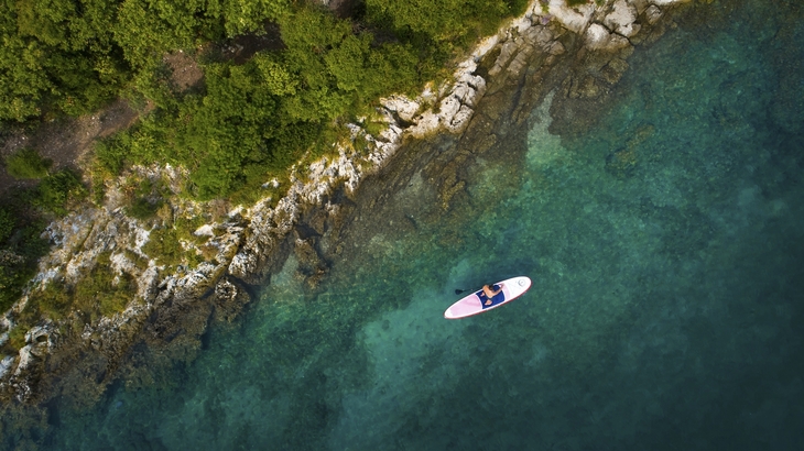 Person auf Paddleboard auf klarem Wasser neben felsiger Küste und bewaldetem Ufer.