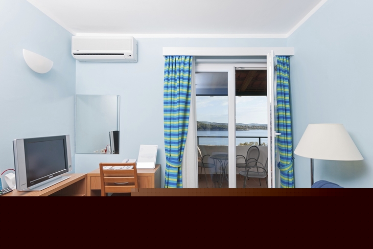 Blick in ein helles Zimmer mit Schreibtisch, TV, Sofa und Zugang zu einem Balkon mit Meerblick.