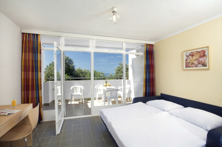 Helles Apartment mit Balkon, Doppelbett, Tisch und Stühlen, Meerblick im Hintergrund.