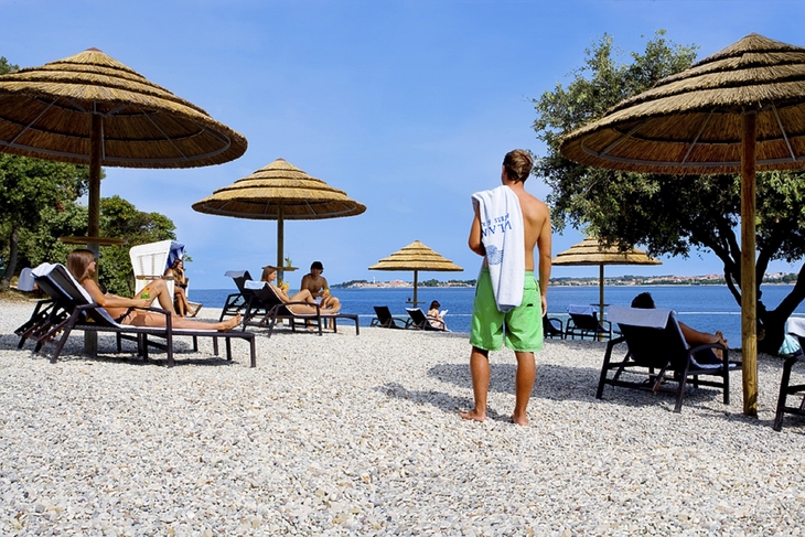 Personen entspannen sich unter Strohdächern am Kiesstrand.