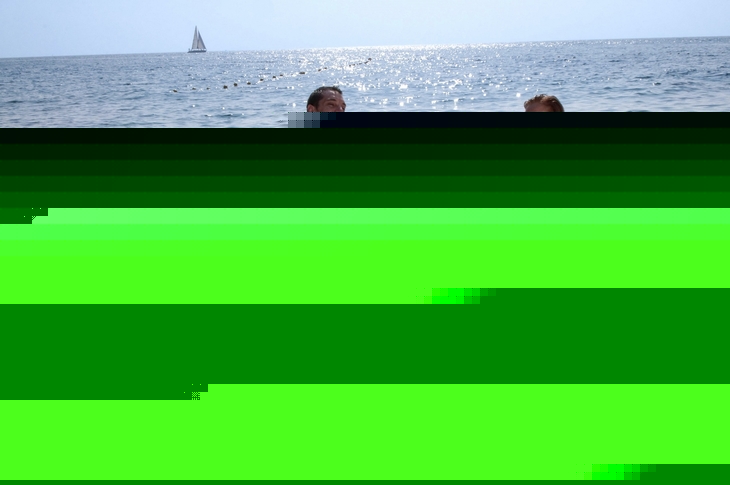 Familie am Strand mit Luftmatratzen im Wasser, im Hintergrund ein Segelboot.
