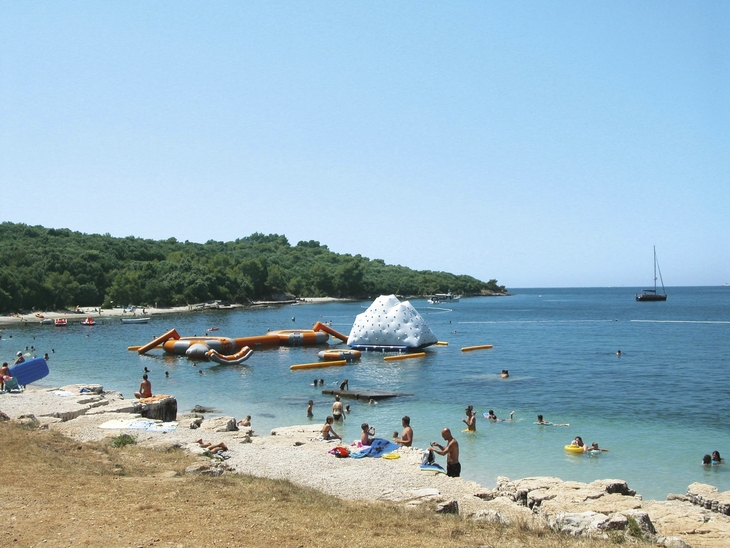 Strand mit Schwimmern und Hindernisparcours im Meer, bewaldete Küste im Hintergrund.